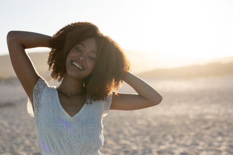 A happy, attractive mixed race woman with hands behind her head, enjoying free time on beach on a sunny day, wearing a white dress with sun shining behind her.